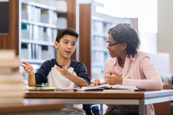 A serious middle school age boy sits at a table with a mature female tutor in his school library.  He gestures as he tells her his issues with the homework.